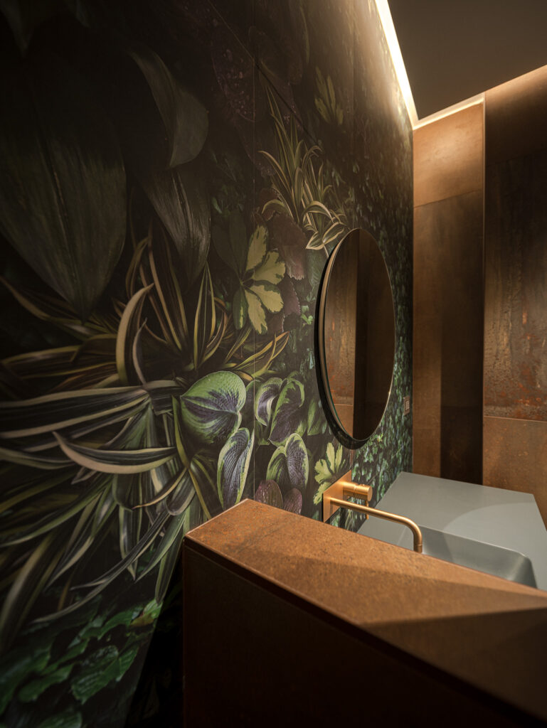 Parete botanica con lavabo grigio, specchio tondo e rubinetto bronzato