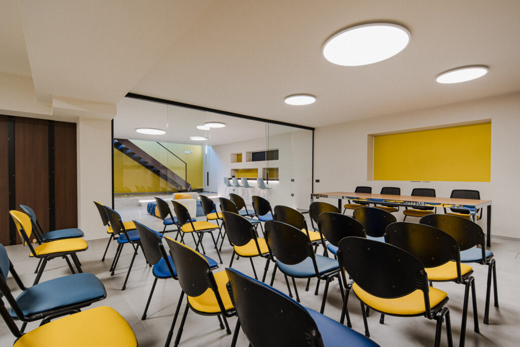 Sala formazione vetrata con sedie colorate e lounge visibile sullo sfondo