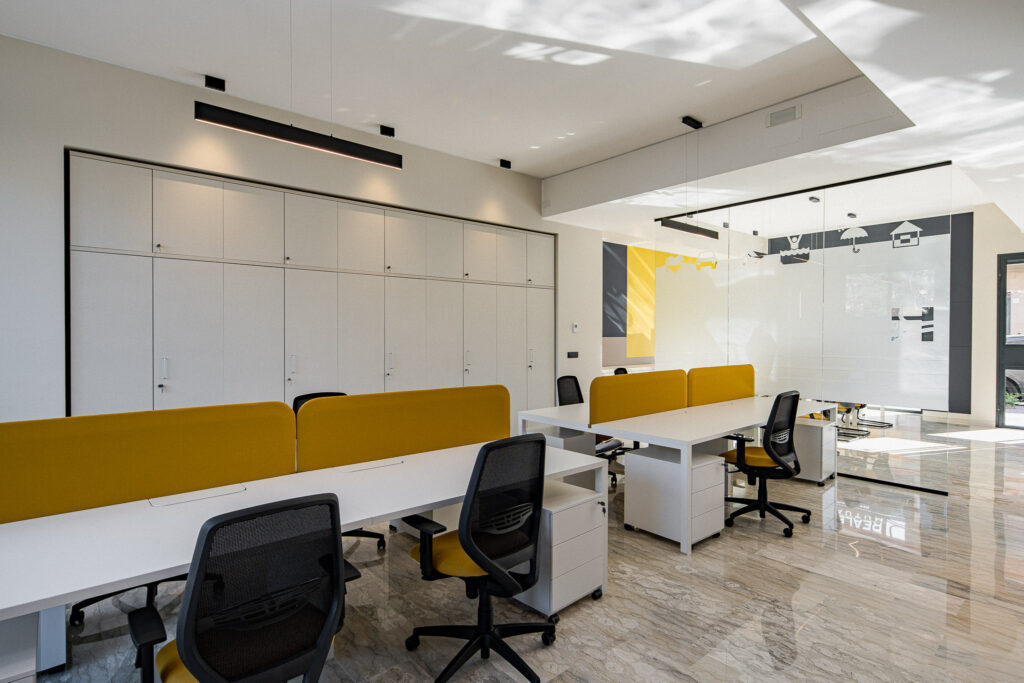 Postazioni da ufficio con pannelli gialli e sala vetrata sullo sfondo