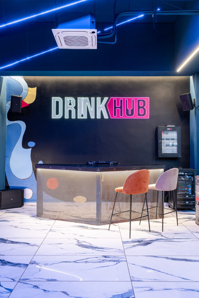 Consolle DJ davanti al logo luminoso Drink Hub con sgabelli colorati