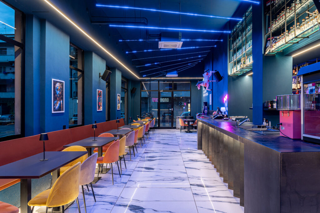 Sala del Drink Hub con bancone laterale, tavoli e ingresso sullo sfondo