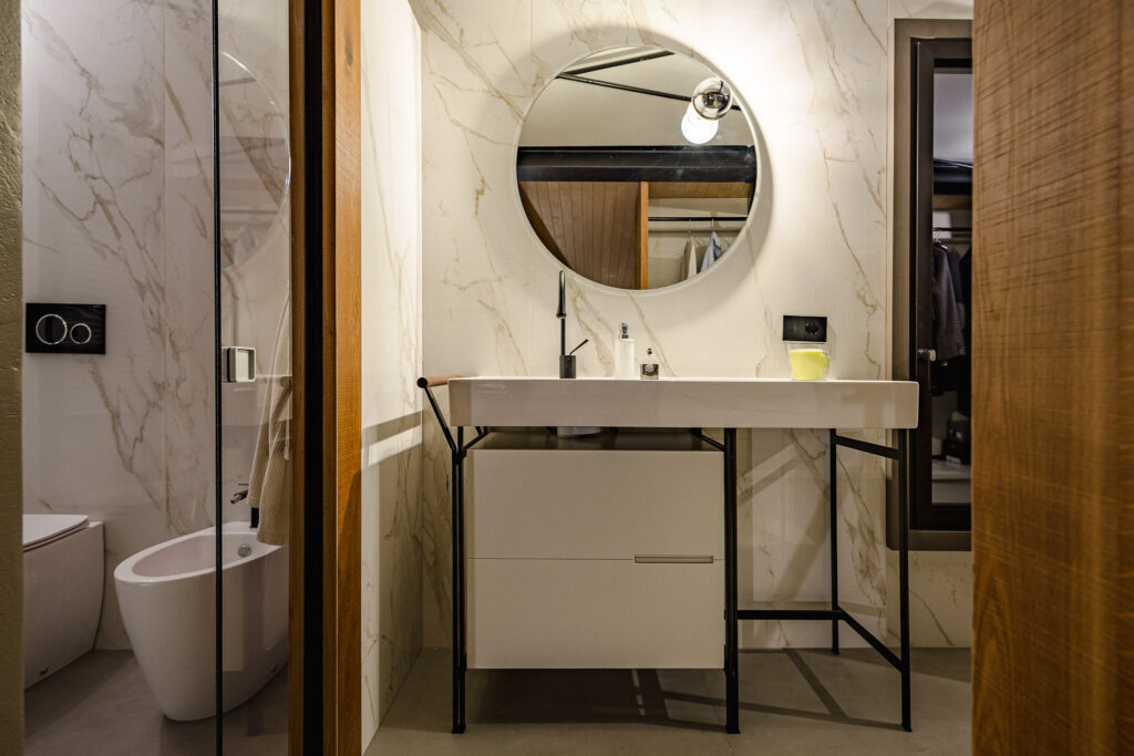 Bagno con lavabo bianco sospeso, specchio rotondo e rivestimento effetto marmo